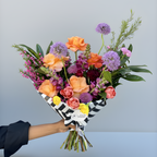 A vibrant floral bouquet held by a person's arm against a clean, light gray studio background. The bouquet features a mix of orange, purple, pink, and red flowers with green foliage, wrapped in a black and white striped paper sleeve with a small white tag that reads 'Flowers Direct'.