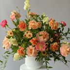 A vibrant floral arrangement featuring peach, pink, and cream roses with green foliage, presented in a white ceramic pot on a white ribbed pedestal against a neutral gray background.