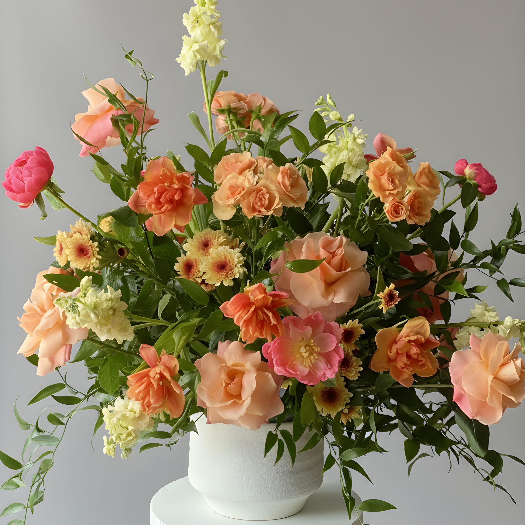 A vibrant floral arrangement featuring peach, pink, and cream roses with green foliage, presented in a white ceramic pot on a white ribbed pedestal against a neutral gray background.