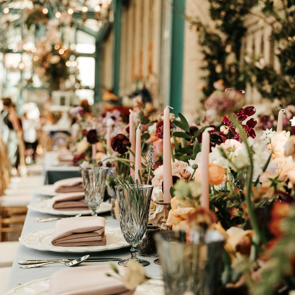 A beautifully styled wedding reception table by Posey Floral with floral centerpieces, pink taper candles, clear cut glass goblets, white plates, and blush pink folded napkins in a warm, softly blurred indoor venue.