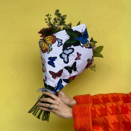Bouquet of flowers wrapped in paper with butterfly design held by a person wearing an orange jacket against a yellow background.