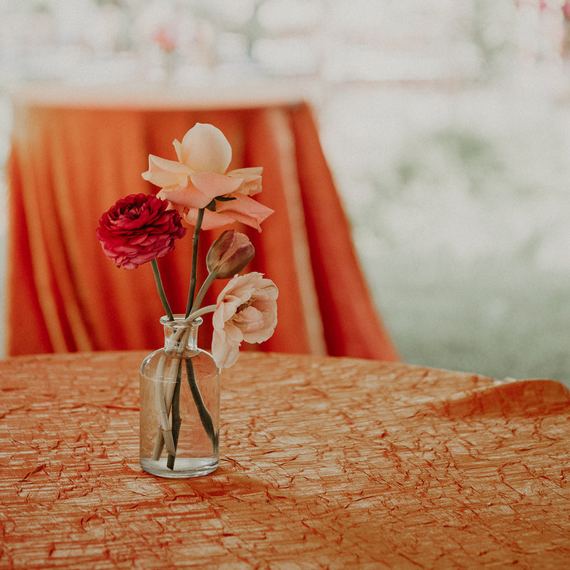 A small clear glass vase holding three flowers, including a peach bloom, a deep red rose, and a pale peach blossom, placed on an orange textured tablecloth with a softly blurred orange draped chair and white wall in the background.