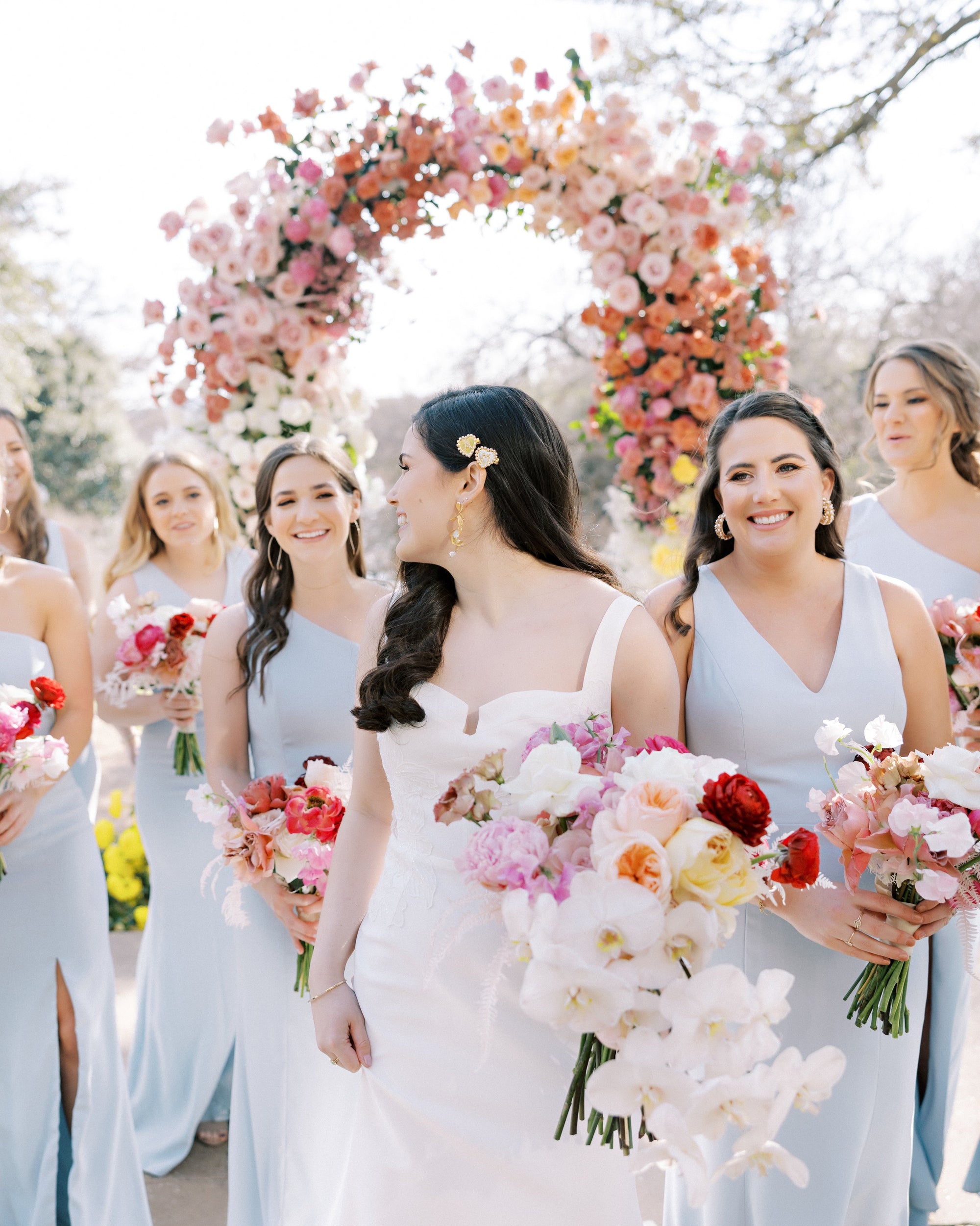 A bride in a white wedding dress walks outdoors with bridesmaids wearing light blue dresses, holding colorful flower bouquets under a large floral arch.