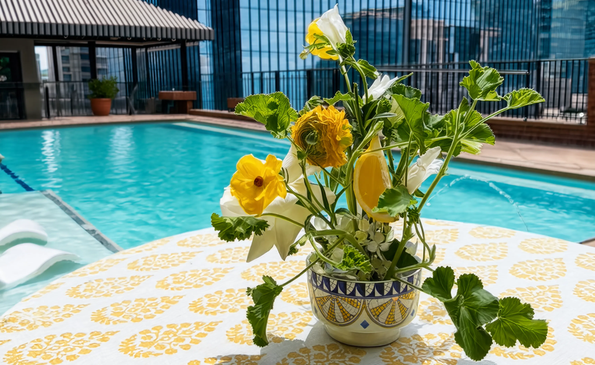 Floral arrangement in a pot on a table with a pool and cityscape in the background