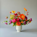 A vibrant, multicolored floral arrangement featuring orange, pink, red, yellow, and purple flowers with green leaves, presented in a textured white ceramic vase against a neutral gray background.