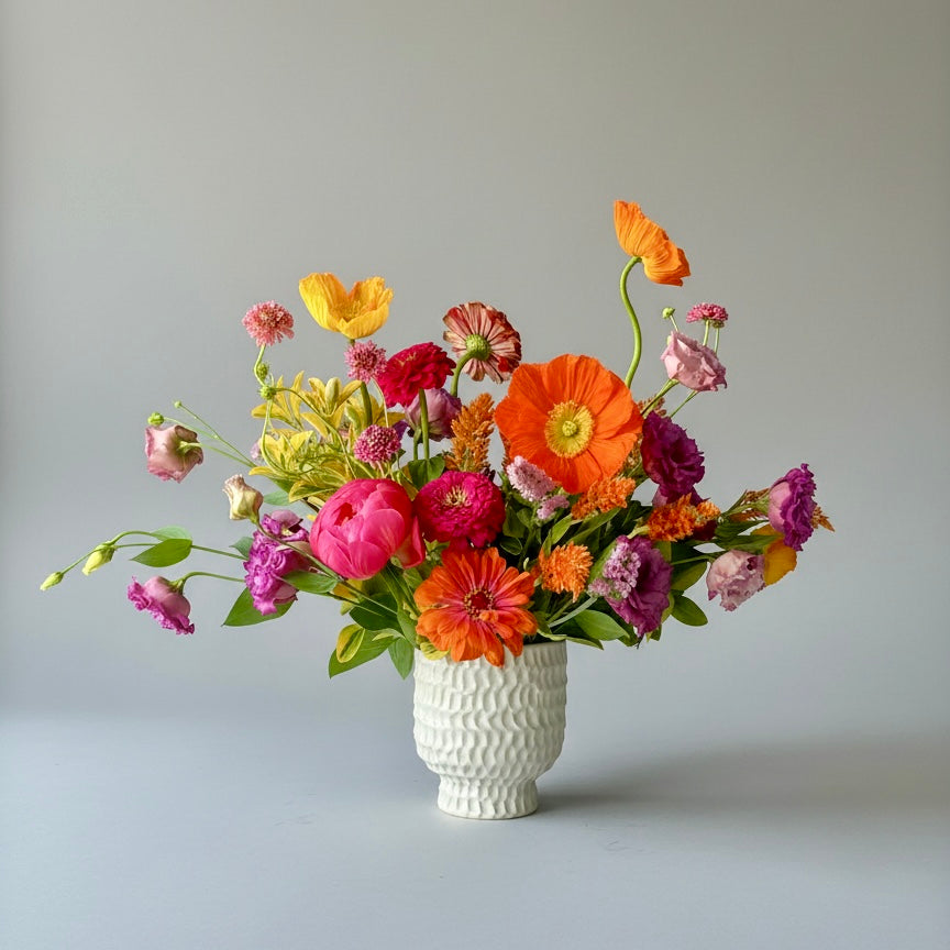 A vibrant, multicolored floral arrangement featuring orange, pink, red, yellow, and purple flowers with green leaves, presented in a textured white ceramic vase against a neutral gray background.