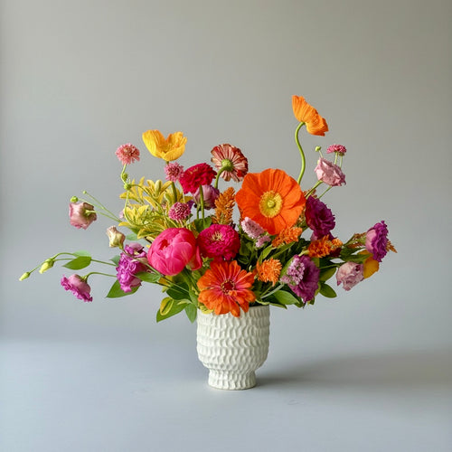 A vibrant, multicolored floral arrangement featuring orange, pink, red, yellow, and purple flowers with green leaves, presented in a textured white ceramic vase against a neutral gray background.