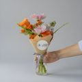 A hand holding a small floral bouquet in a clear glass jar vase against a plain light gray background. The bouquet includes orange roses, pink daisies, and small purple flowers, wrapped in tan paper with a round white label reading 'little posy'.