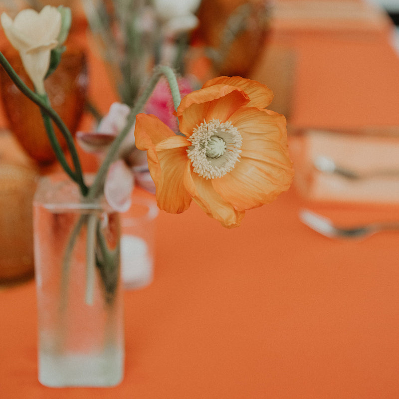 A close-up of a bright orange poppy flower in a clear glass vase on an orange table runner, with blurred flowers and tableware in the background.