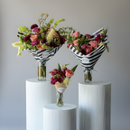 Three floral arrangements in clear glass vases displayed on white cylindrical pedestals against a light gray studio background. Two bouquets are wrapped in black-and-white striped paper, and one smaller bouquet is wrapped in tan paper.