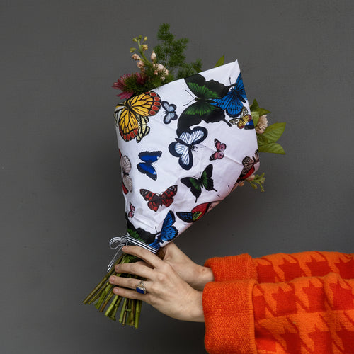 Bouquet of flowers wrapped in paper with butterfly design held by a person wearing an orange jacket against a yellow background. austin tx