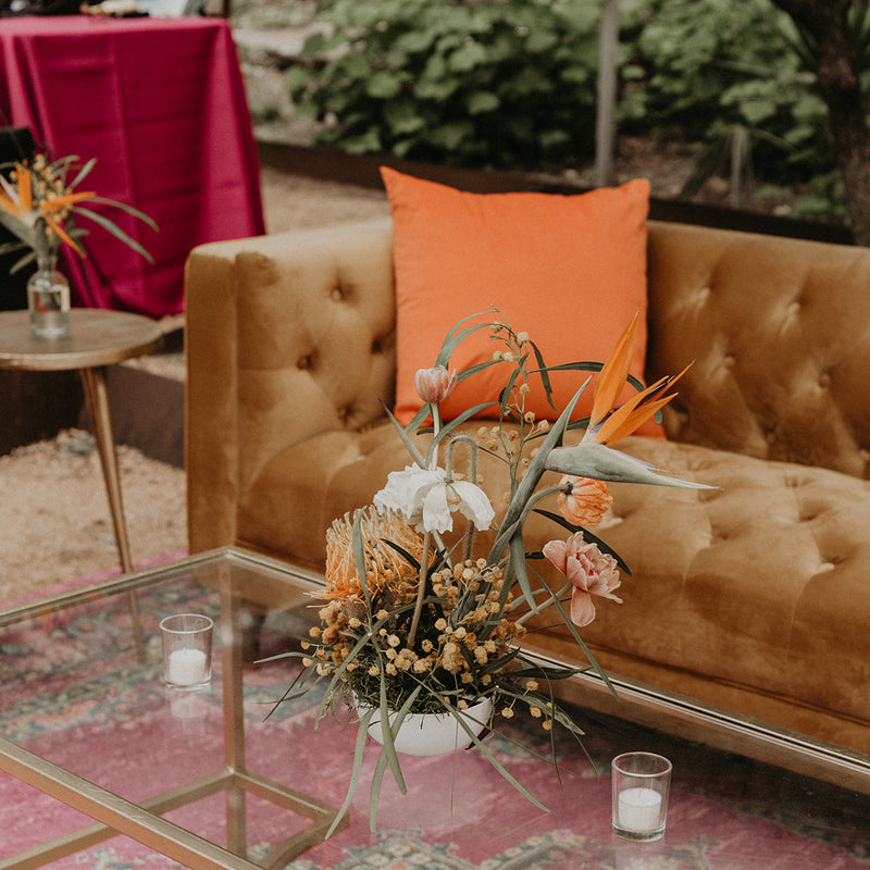 A styled outdoor lounge scene with a tan tufted velvet sofa, a bright orange accent pillow, a glass-top coffee table with a gold frame, and a floral arrangement on a pink patterned rug.