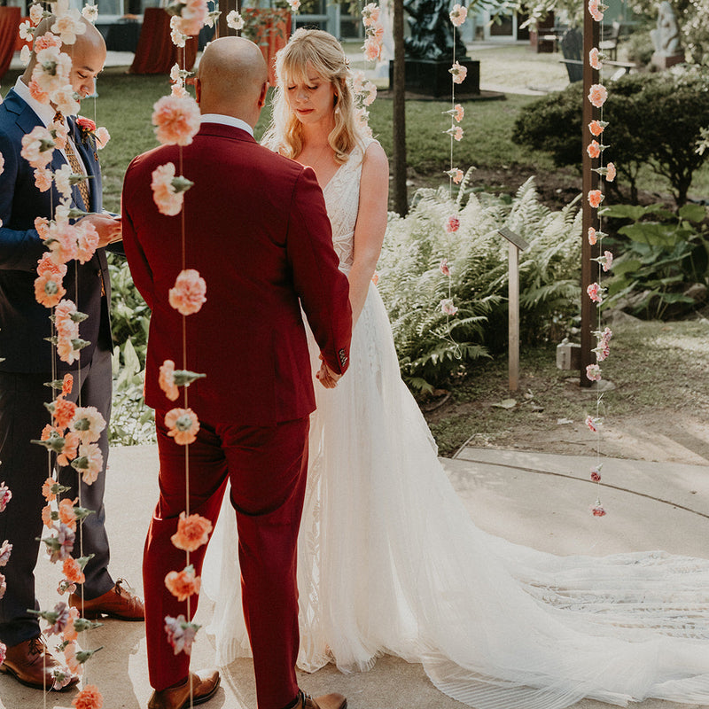 A bride in a white wedding gown and a groom in a deep burgundy suit stand under a floral archway during an outdoor wedding ceremony. The scene is decorated with hanging pink and peach flowers, set under a wooden pavilion with trees in the background.
