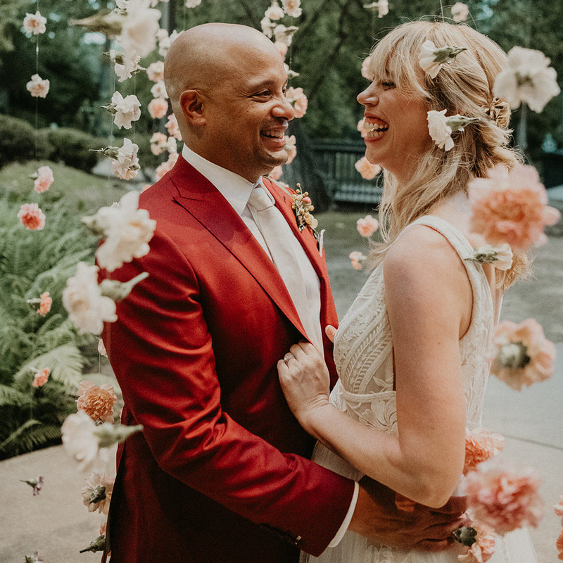 A smiling couple embracing under hanging floral decorations. The man wears a bright red suit jacket, white shirt, and red tie, while the woman wears a sleeveless cream lace wedding dress with a floral hair accessory.