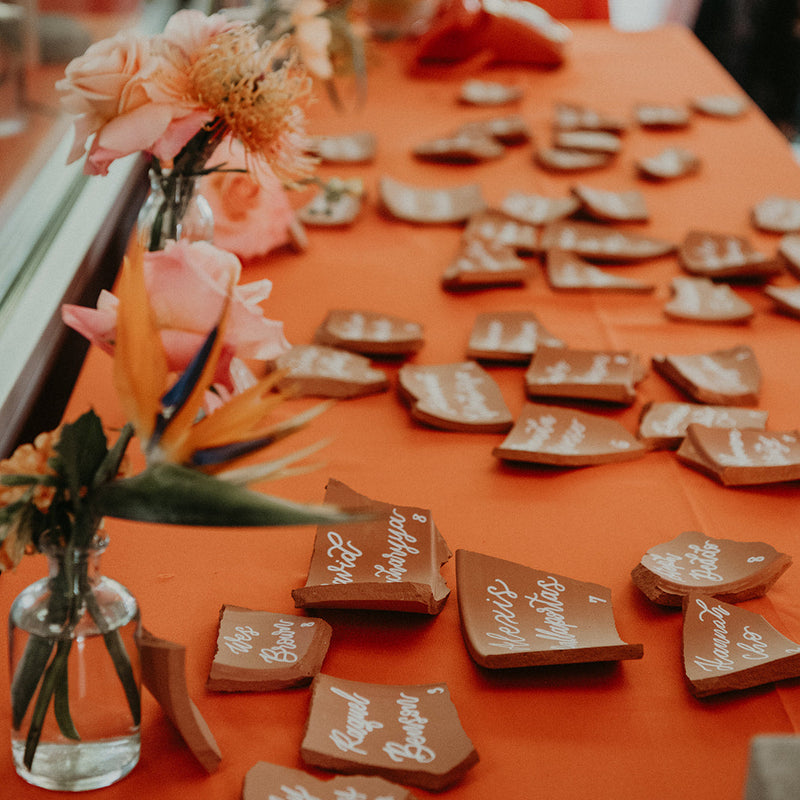 A long table covered with a bright orange cloth, scattered with small brown broken-glass style place cards or name tags, decorated with floral arrangements in glass vases.