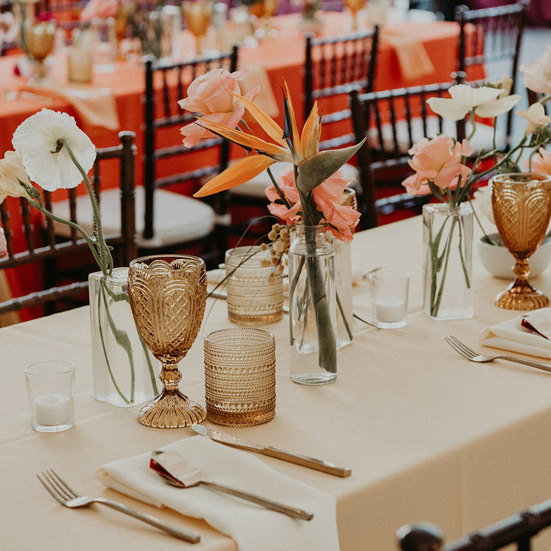 A styled wedding reception table setting with a cream tablecloth, gold goblets, clear glass candle holders, and floral centerpieces with peach and white flowers, with blurred chairs and guests in the background.