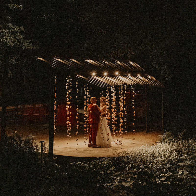 A couple stands under a small illuminated canopy in a dark outdoor nighttime setting, surrounded by trees and string lights. The bride wears a white gown and the groom wears a dark formal outfit.